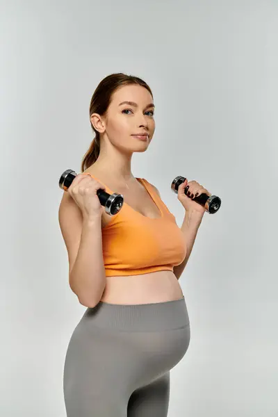 Eine junge, sportliche Schwangere in aktiver Kleidung hält vor grauem Hintergrund zwei Kurzhanteln in den Händen. — Stockfoto