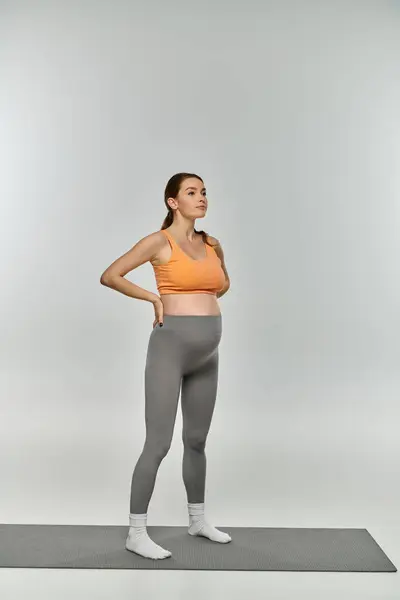 Una mujer embarazada deportiva en traje de entrenamiento se para con confianza en una esterilla con las manos en las caderas, mostrando su fuerza y vitalidad. - foto de stock