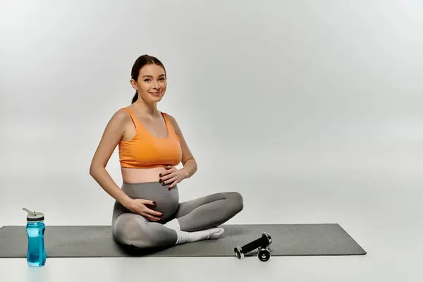A pregnant woman in active wear sits on a yoga mat near a bottle of water. — Stock Photo