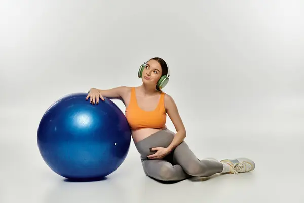A sporty pregnant woman in active wear sitting on the floor next to a large blue ball, deep in thought. — Stock Photo