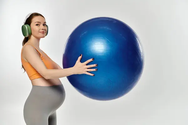 A young athletic pregnant woman in activewear balances a large blue ball in her hands on a grey background. — Stock Photo