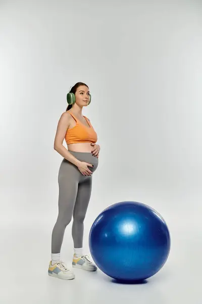 Eine junge, sportliche Schwangere in aktiver Kleidung steht anmutig neben einem blauen Trainingsball auf grauem Hintergrund.. — Stockfoto