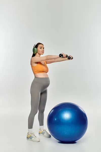 Sportliche Schwangere in aktiver Kleidung beim Training auf dem Gymnastikball mit Kopfhörern auf grauem Hintergrund. — Stockfoto