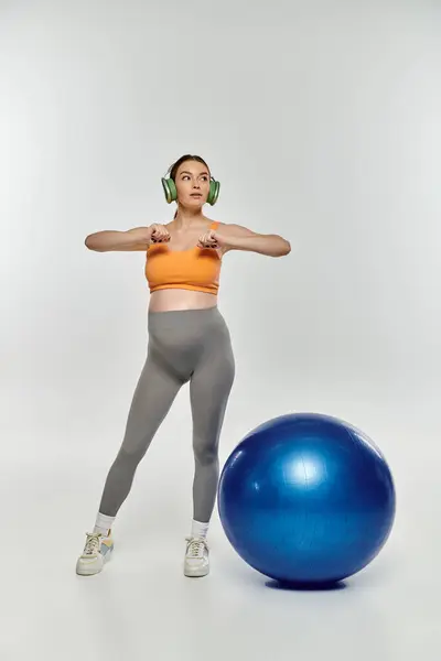 A pregnant woman in activewear stands next to a bright blue exercise ball on a grey background. — Stock Photo