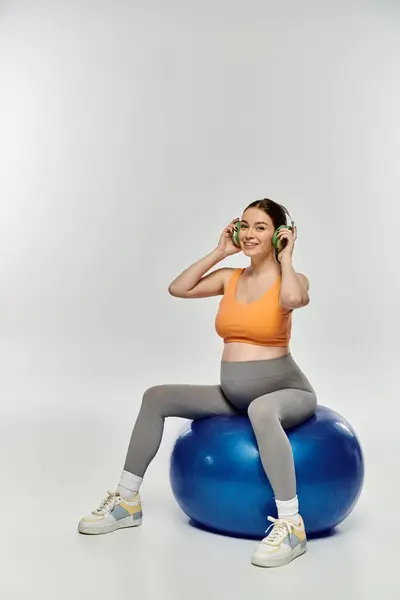 Junge schwangere Frau balanciert in aktiver Kleidung auf blauem Ball, tief in der Musik verstrickt. — Stockfoto