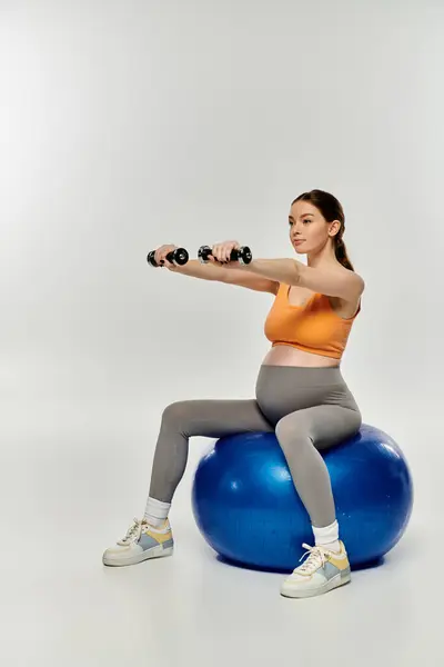 Une femme enceinte en tenue de sport s'assoit sur une balle de fitness, soulevant un haltère dans une pose gracieuse et équilibrée. — Photo de stock