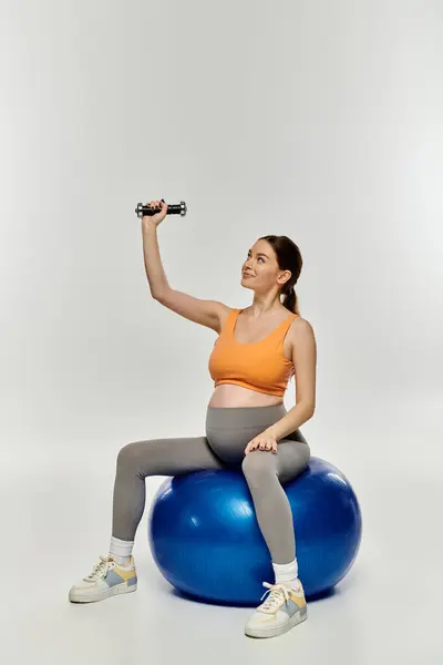 Eine schwangere Frau in Aktivkleidung demonstriert Gleichgewicht, indem sie mit einer Hantel auf einem Ball sitzt. — Stockfoto