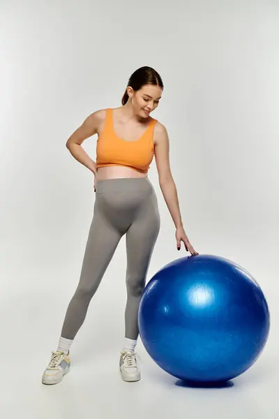 A young, sporty pregnant woman in active wear exercises next to a fitness ball on a grey background. — Stock Photo
