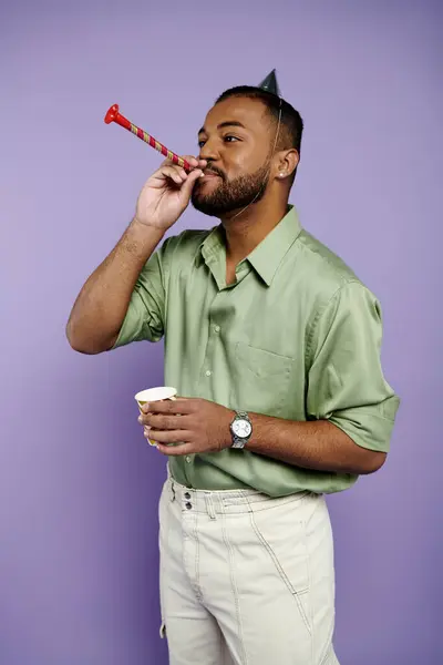 Ein fröhlicher junger afroamerikanischer Mann mit Geburtstagshut bläst Party-Horn vor violettem Hintergrund. — Stock Photo