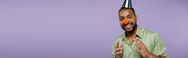 A young African American man with braces on a purple background is happily balancing a clown nose on top of his head. — Stock Photo