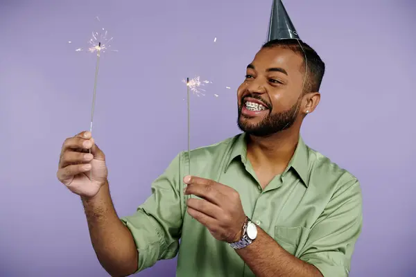 A young African American man in braces joyfully holding sparklers while wearing a party hat on a purple background. — Stock Photo