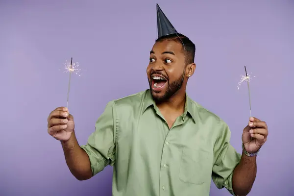 Young African American man with braces, smiling, holds two sparklers in party hat against vibrant purple background. — Stock Photo