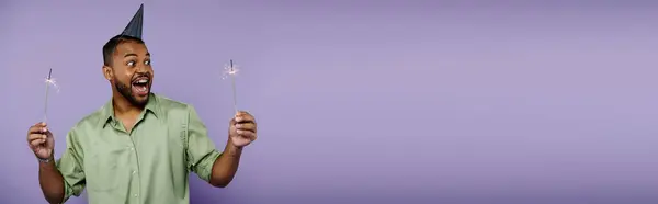 Young African American male with braces balancing two sparklers, wearing a festive party hat on a purple background. — Stock Photo