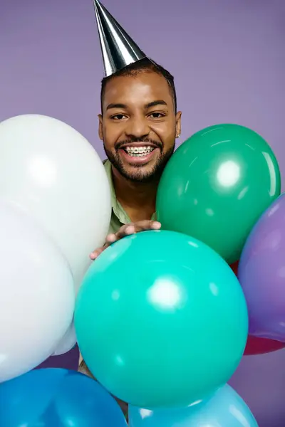 Junger Afroamerikaner mit Zahnspange hält fröhlich bunte Luftballons mit festlichem Partyhut vor lila Hintergrund. — Stockfoto