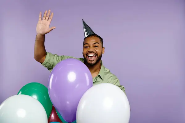 Young African American man in party hat smiling, holding colorful balloons against a purple background. — Stock Photo