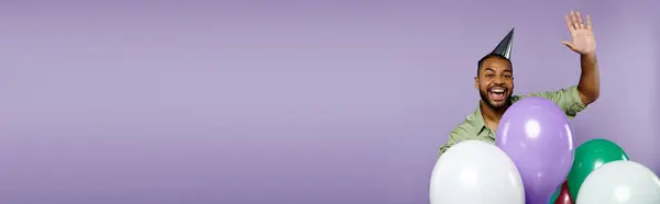 Young African American man wearing braces enthusiastically holds balloons in a party hat against a purple backdrop. — Stock Photo