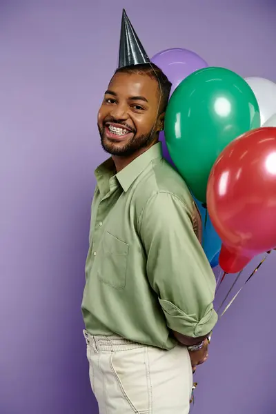 Young African American man in a party hat happily holds colorful balloons on a purple background. — Stock Photo