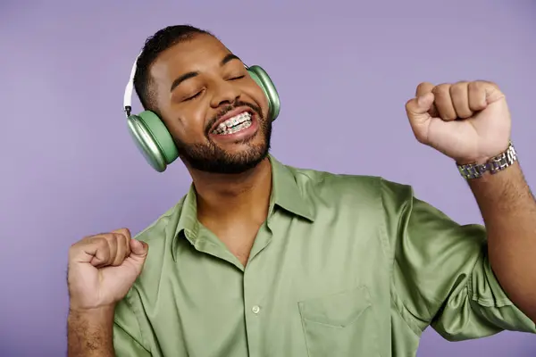 Glücklicher junger afroamerikanischer Mann mit Hosenträgern in grünem Hemd und Kopfhörer auf lila Hintergrund. — Stockfoto