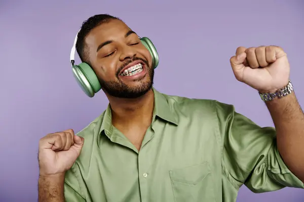 Happy young African American man with braces wearing a green shirt and headphones on a purple background. — Stock Photo