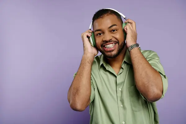 Un jeune homme afro-américain heureux avec des bretelles sourit largement pour la caméra tout en portant des écouteurs sur un fond violet vibrant. — Photo de stock