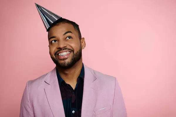 Smiling young African American man in braces wearing a party hat on a pink background. — Stock Photo