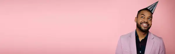 Young African American man, adorned in braces, exudes happiness while showcasing a vibrant party hat on a pink background. — Stock Photo