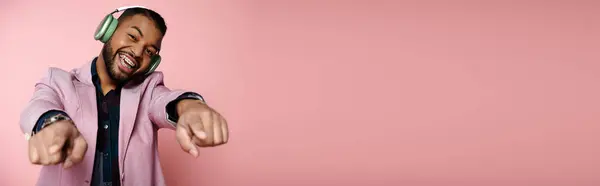 Young African American man joyfully points while wearing headphones against a vibrant pink backdrop. — Stock Photo
