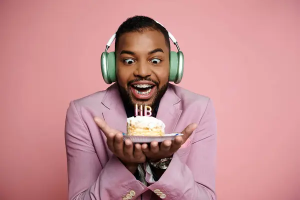 Young African American man with braces happily holds a piece of cake while wearing headphones on a pink background. — Stock Photo