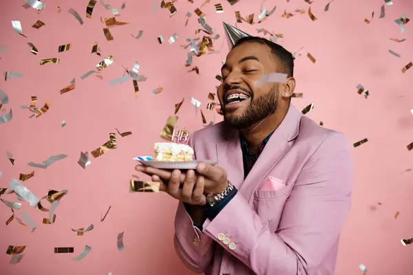 Junger Afroamerikaner im rosafarbenen Anzug hält fröhlich eine Torte und Konfetti auf einem leuchtend rosafarbenen Hintergrund. — Stockfoto