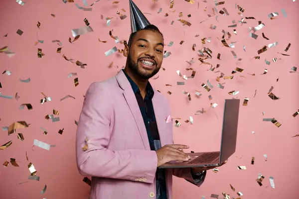 Joyful young African American man in braces balancing a laptop, dressed in a party hat on a vibrant pink background. — Stock Photo