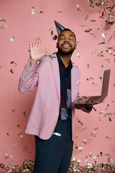 Ein junger Afroamerikaner in Hosenträgern lächelt, während er einen Laptop in der Hand hält und einen Partyhut auf rosa Hintergrund trägt. — Stockfoto