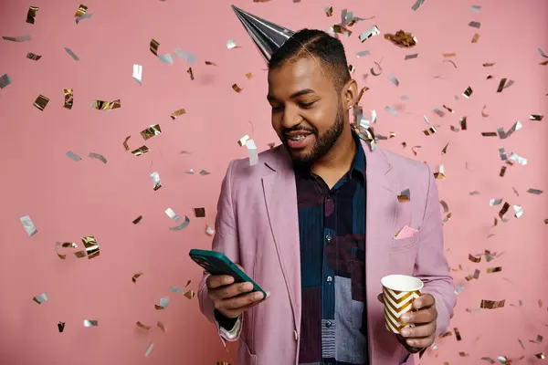 Young African American man in braces, happy, holding cup and cell phone, standing in party hat on a pink background. — Stock Photo