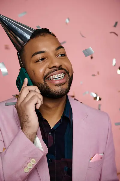 Молодой, счастливый афроамериканец в брекетах, в шляпе для вечеринок, разговаривает по мобильному телефону на розовом фоне. — стоковое фото