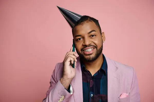 Joven hombre afroamericano con frenos felizmente habla en el teléfono celular mientras usa un sombrero de fiesta festiva contra un telón de fondo rosa. - foto de stock