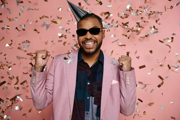 Jeune homme afro-américain rayonne dans un chapeau de fête et des lunettes de soleil sur un fond rose vif. — Photo de stock