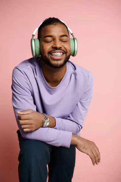 Young African American man happily listening to music on headphones while seated on a stool against a pink background. — Stock Photo