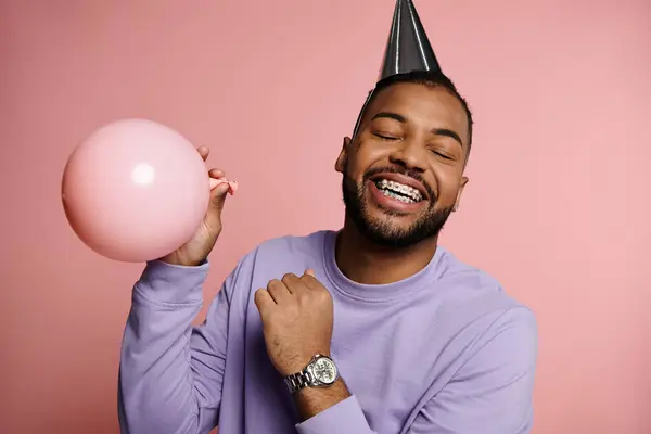 Giovane uomo afroamericano con bretelle sorridenti e con in mano un palloncino, indossa un colorato cappello da festa su sfondo rosa. — Foto stock