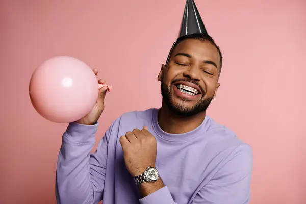 Young African American man with braces smiling and holding a balloon, wearing a colorful party hat on a pink background. — Stock Photo