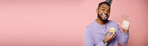 Щасливий, молодий афроамериканський чоловік з брекетами, у фіолетовій сорочці, смакує випічку на рожевому фоні. — стокове фото