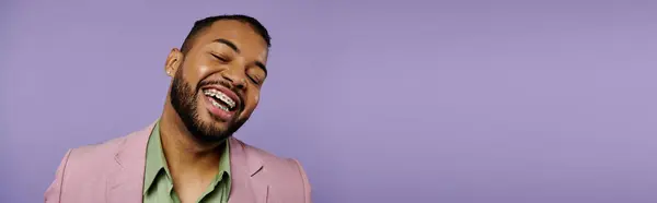 Junger afroamerikanischer Mann mit Zahnspange lacht fröhlich in einer stylischen rosafarbenen Jacke vor einer lebhaften lila Kulisse. — Stockfoto