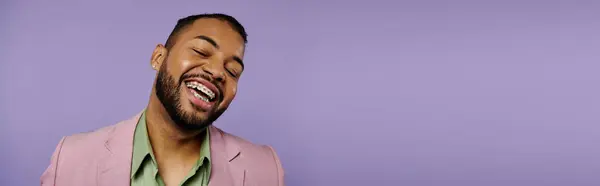 Young African American man with braces laughing joyfully in a stylish pink jacket against a vibrant purple backdrop. — Stock Photo