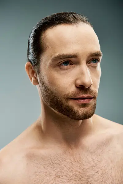 Un hombre sin camisa que emana masculinidad y confianza, mostrando su barba rugosa en un estudio sobre un fondo gris. - foto de stock