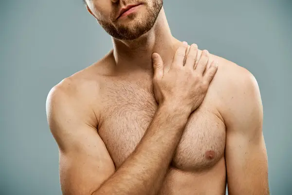 Un hombre rugoso y sin camisa con barba se levanta confiadamente con las manos en el pecho sobre un fondo gris del estudio. - foto de stock