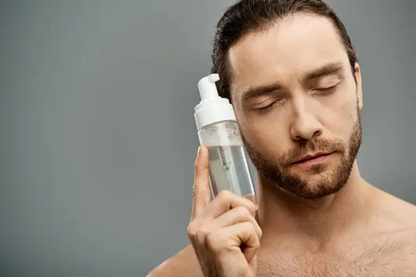Ein hemdloser, bärtiger Mann hält vor grauer Studiokulisse selbstbewusst eine Flasche Reinigungsmittel in der Hand. — Stockfoto