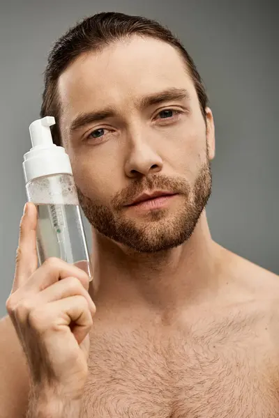 Ein Mann mit Bart hält eine Flasche mit Reinigungsmittel in einem Studio vor grauem Hintergrund. — Stockfoto
