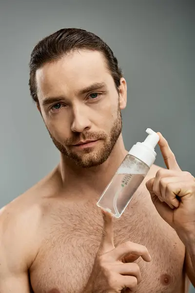 Un homme torse nu et beau avec une barbe tient gracieusement une bouteille de nettoyant sur fond de studio gris. — Photo de stock