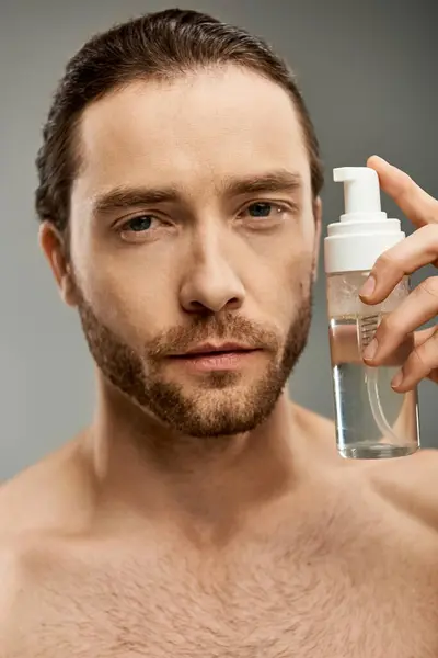A shirtless, bearded man gracefully holds a bottle of foam cleanser against a grey backdrop. — Stock Photo