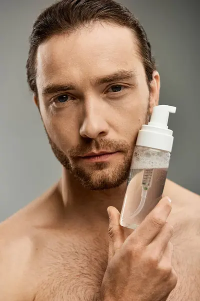 Ein gutaussehender, hemdloser Mann mit Bart und einer Flasche Reinigungsmittel vor grauem Hintergrund. — Stockfoto