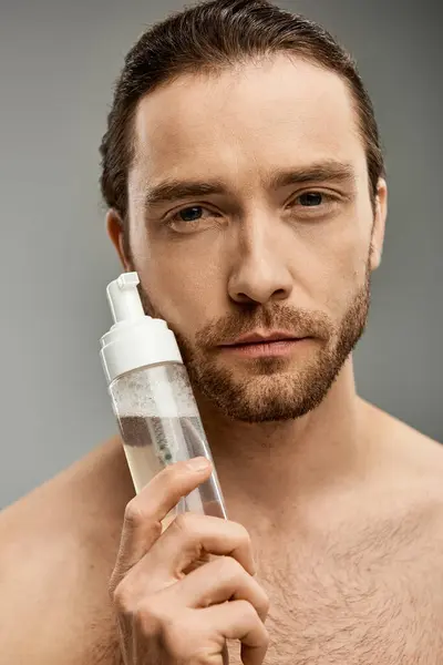A shirtless, bearded man holding a bottle of cleanser in a studio against a grey background. — Stock Photo