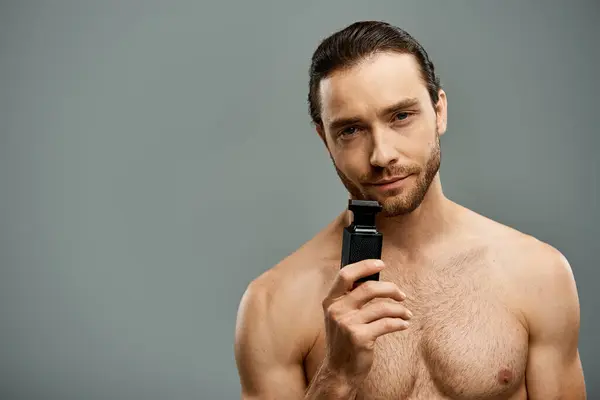 Hemdloser Mann mit Bart hält eine Parfümflasche vor grauer Studiokulisse. — Stockfoto